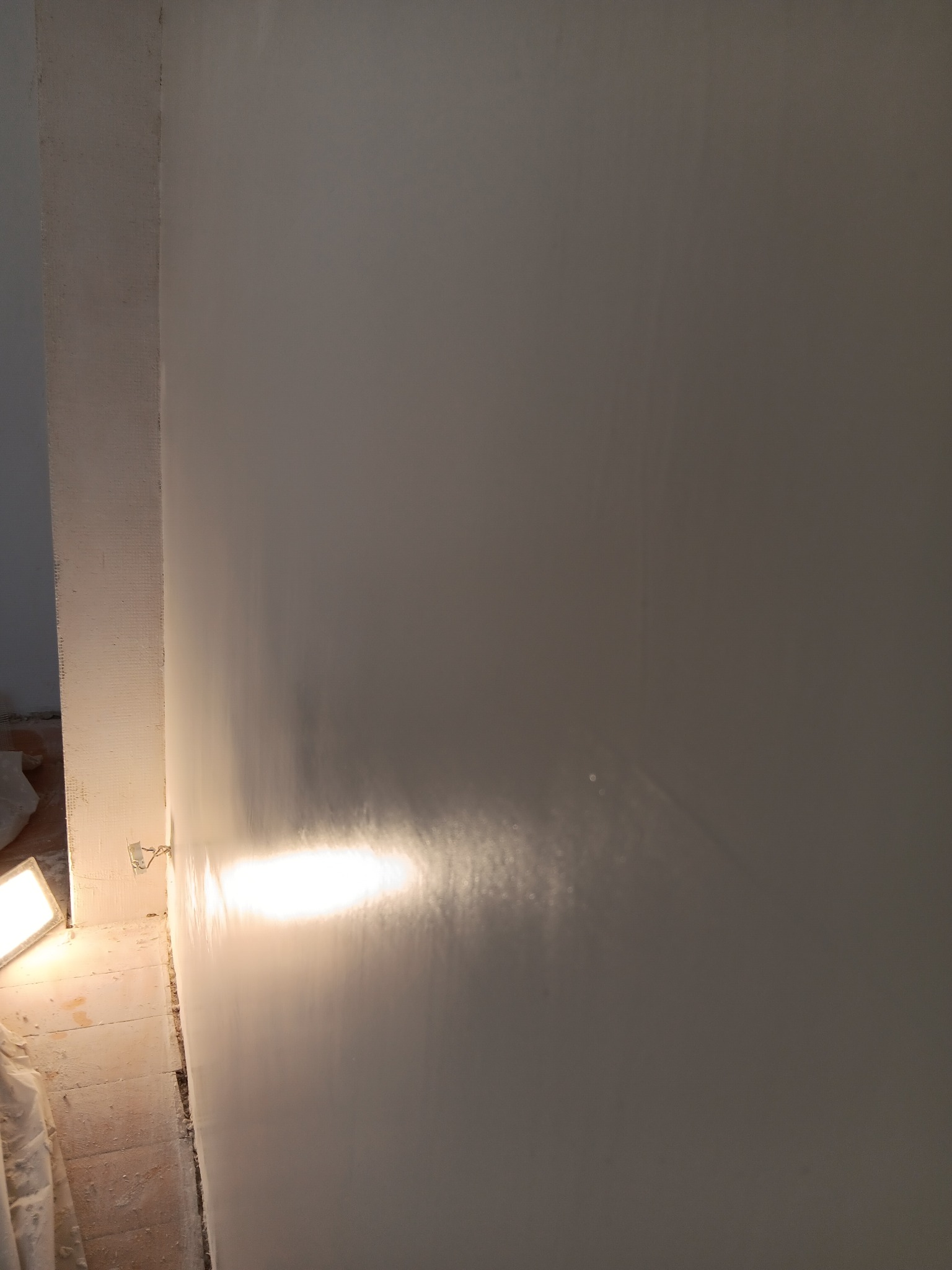 Świeżo nałożona, gładka, biała gładź na ścianie, oświetlona lampą budowlaną. Widoczne ślady po szpachlowaniu przy podłodze.