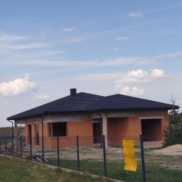 Pokrycia dachowe Andrzej Pierzakowski - Dom jednorodzinny w trakcie budowy z ciemnym dachem z blachodachówki, widoczny zza ogrodzenia panelowego na tle błękitnego nieba z chmurami.