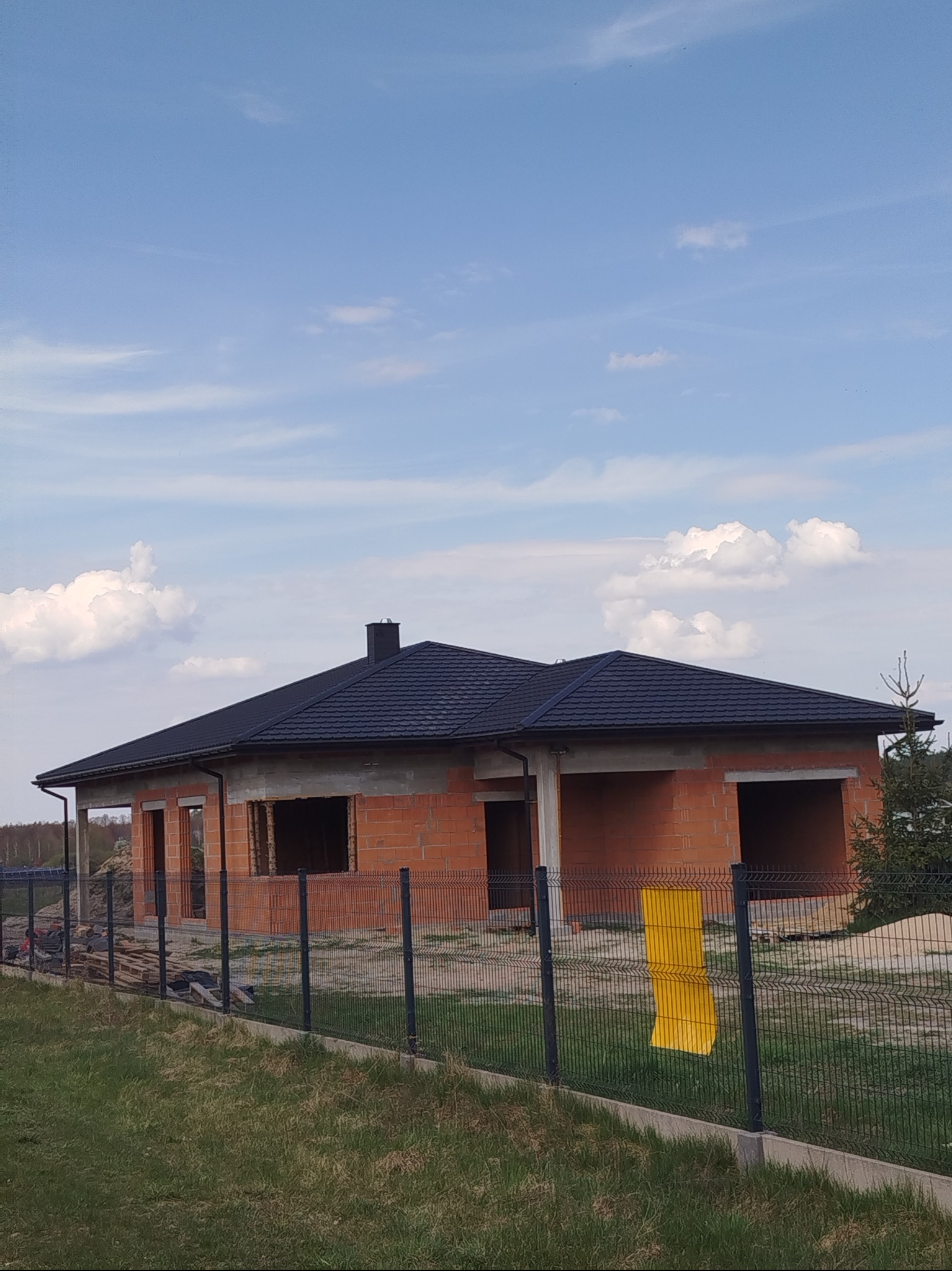 Dom jednorodzinny w trakcie budowy z ciemnym dachem z blachodachówki, widoczny zza ogrodzenia panelowego na tle błękitnego nieba z chmurami.