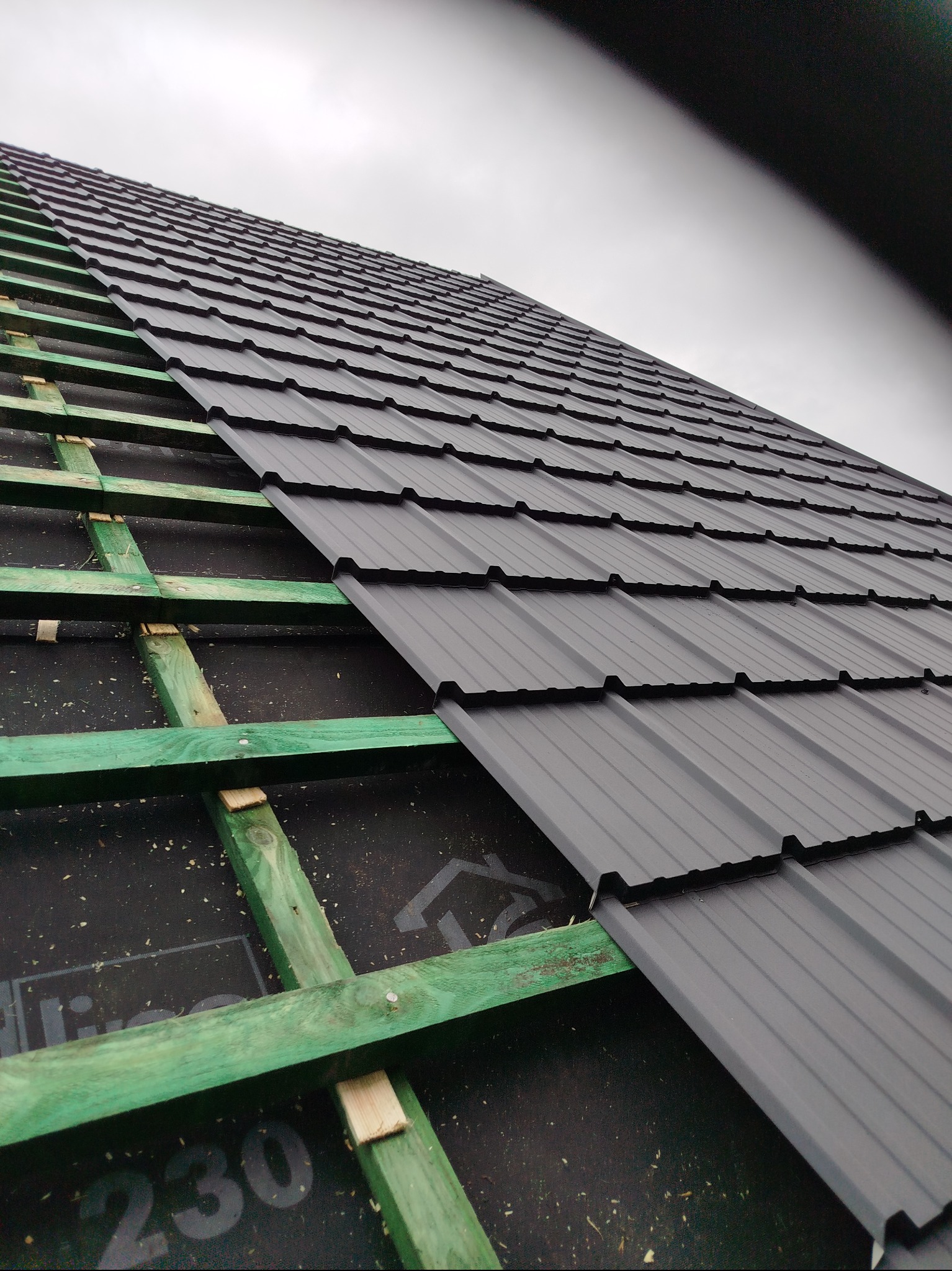 Fragment dachu z szarej blachy trapezowej, widoczna zielona konstrukcja drewniana i czarna membrana dachowa. Ujęcie pod kątem, pochmurne niebo w tle.