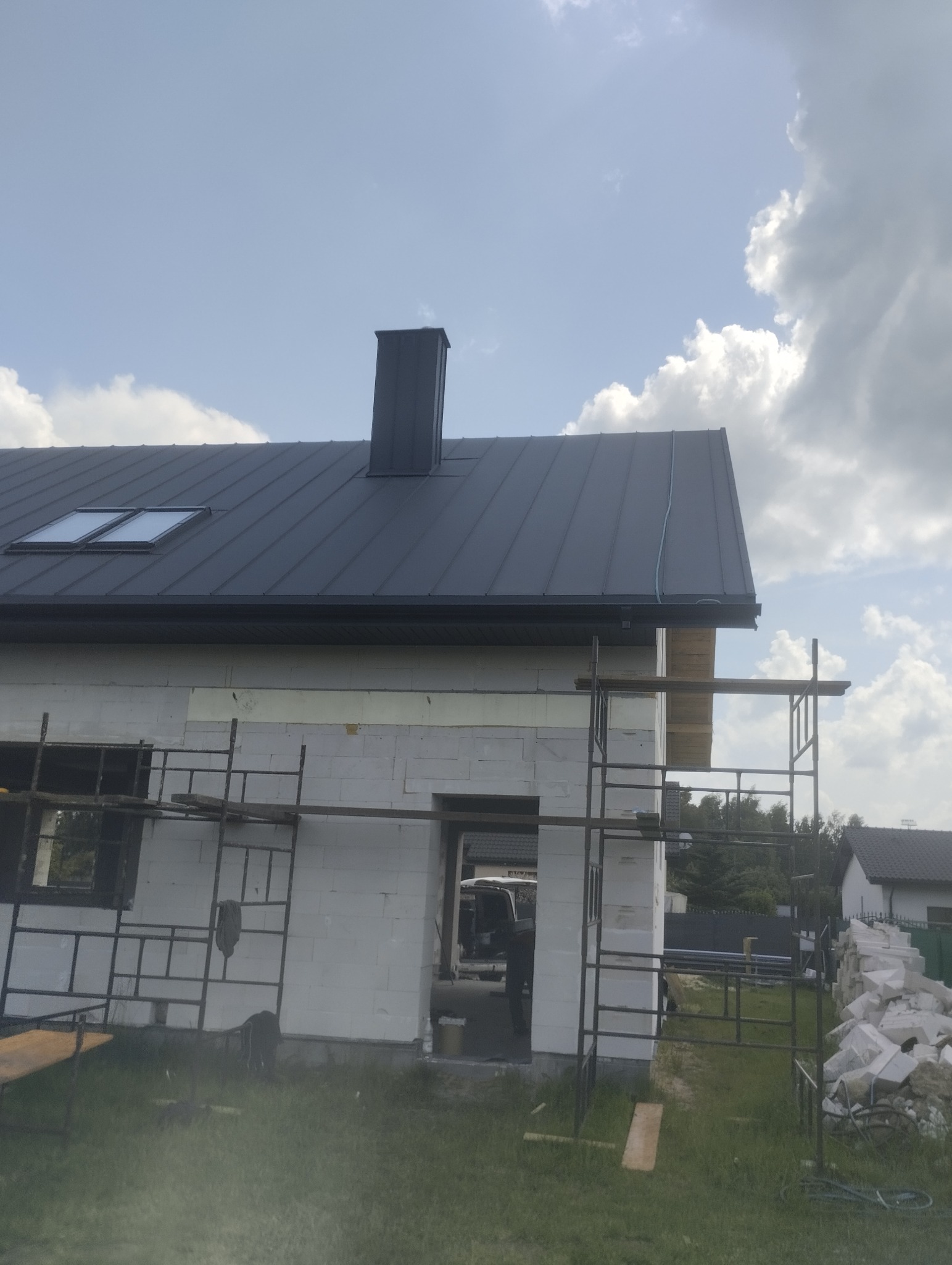 Dom w trakcie budowy z ciemnym dachem z blachy, widoczne rusztowania i komin. Dach z oknami połaciowymi, na tle pochmurnego nieba.