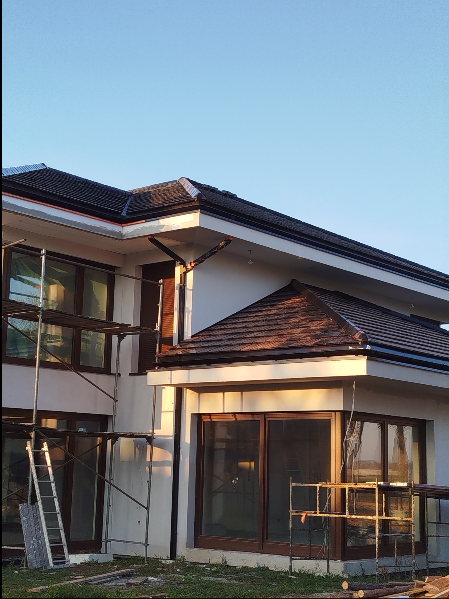 Dwukondygnacyjny dom z brązowym dachem w trakcie budowy, widoczne rusztowania i drabina. Ujęcie w słoneczny dzień.