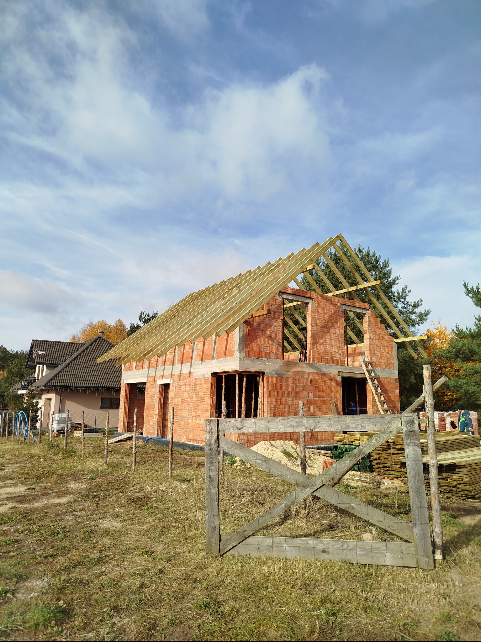 Surowy stan budynku z czerwonej cegły z drewnianą konstrukcją dachu w trakcie budowy, widok z zewnątrz na tle błękitnego nieba i drzew.