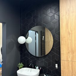 KarolH. - Nowoczesna łazienka z czarnymi, geometrycznymi płytkami 3D, okrągłym lustrem i białą umywalką nablatową. Drewniane szafki dodają ciepła. Minimalistyczne oświetlenie.