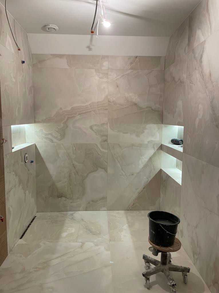 Wykończona łazienka z beżowymi płytkami imitującymi marmur, z podświetlanymi wnękami. Na pierwszym planie wiadro na stołku na kółkach. Widoczne wystające kable.