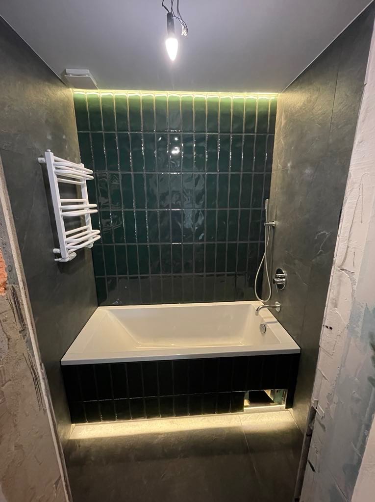 Wykończona łazienka z wanną obudowaną czarnymi płytkami, zielone płytki na ścianie, oświetlenie LED pod wanną i nad płytkami, grzejnik drabinkowy.