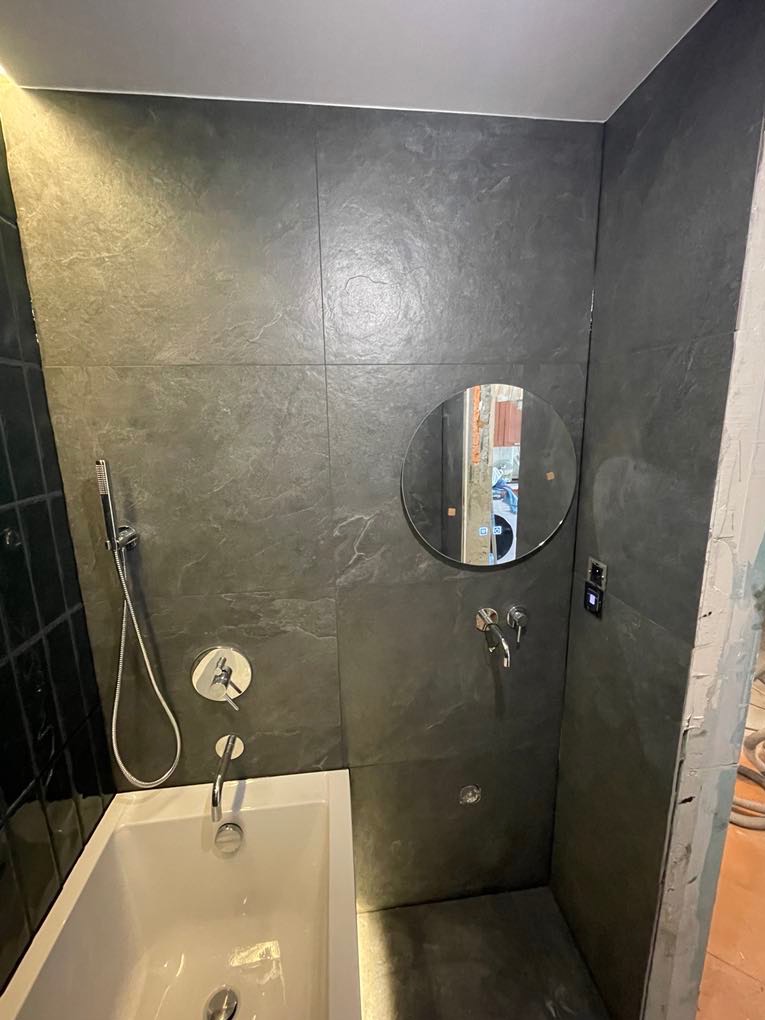 Nowoczesna łazienka z grafitowymi płytkami imitującymi kamień, okrągłym lustrem i białą wanną. Widoczne baterie i słuchawka prysznicowa.