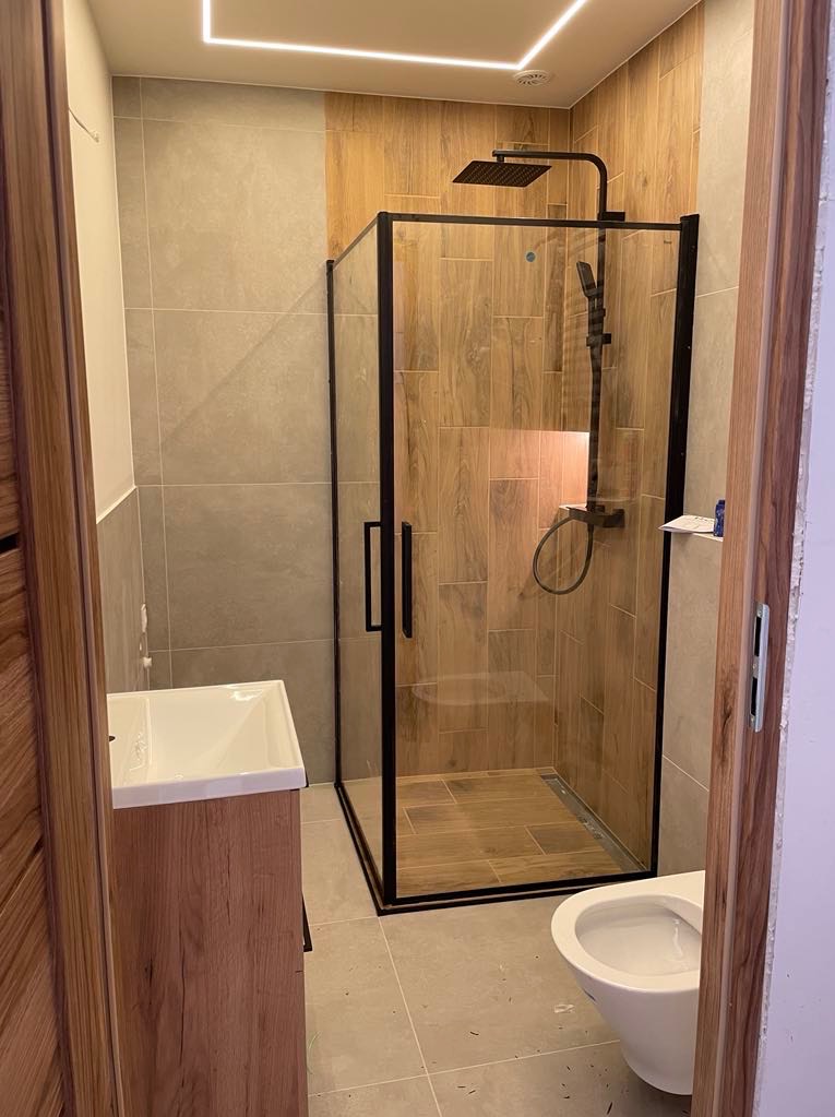 Nowoczesna łazienka z kabiną prysznicową typu walk-in z czarnymi profilami, drewnopodobnymi płytkami i umywalką z szafką w odcieniach drewna. Oświetlenie LED.