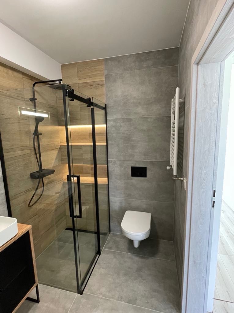 Nowoczesna łazienka z kabiną prysznicową typu walk-in z czarnymi ramami, szarymi płytkami i drewnopodobnymi akcentami. Widoczny grzejnik drabinkowy i toaleta.