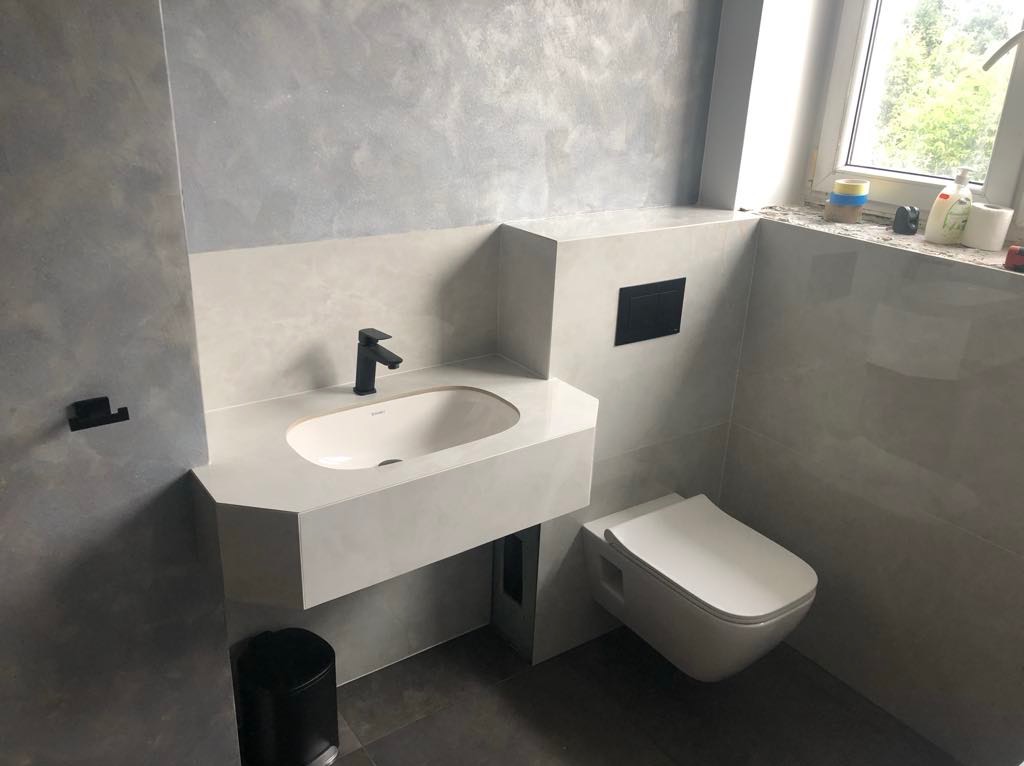 Nowoczesna łazienka po remoncie: szare ściany z efektem betonu, podwieszana toaleta i umywalka z czarną armaturą. Minimalistyczny design, widok na okno.