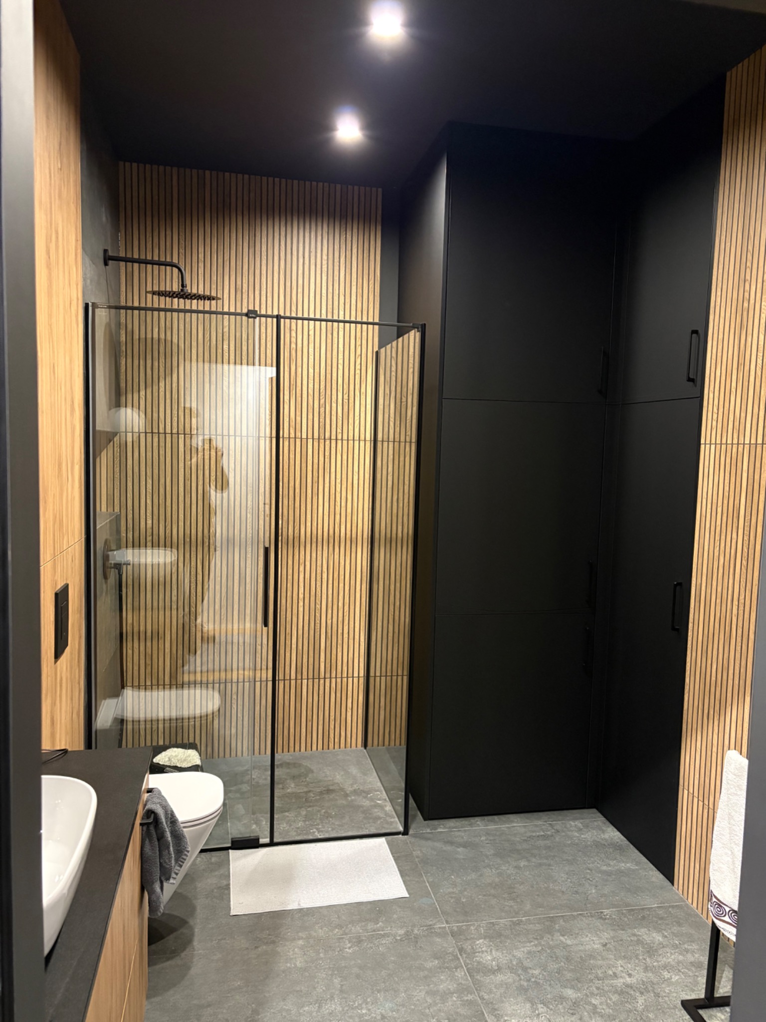 Nowoczesna łazienka z kabiną prysznicową typu walk-in, drewnianymi panelami ściennymi i czarnymi szafkami. Minimalistyczny design z szarą podłogą i białymi elementami ceramiki.