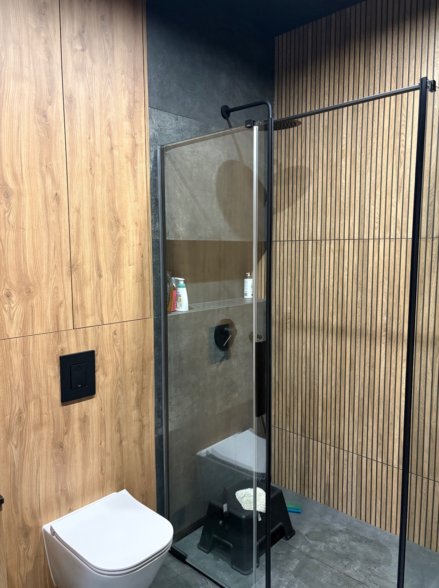 Nowoczesna łazienka z kabiną prysznicową typu walk-in, drewnianymi panelami ściennymi i wiszącą toaletą. Minimalistyczny design z akcentami drewna i szarości.