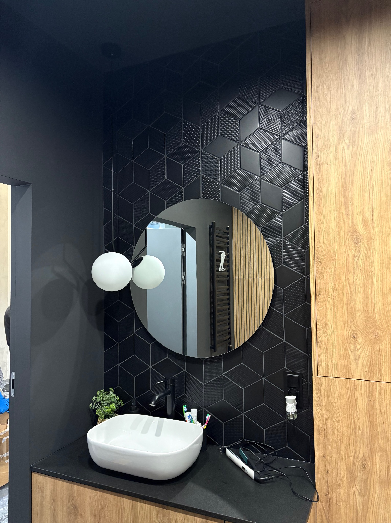 Nowoczesna łazienka z czarnymi, geometrycznymi płytkami 3D, okrągłym lustrem i białą umywalką nablatową. Drewniane szafki dodają ciepła. Minimalistyczne oświetlenie.
