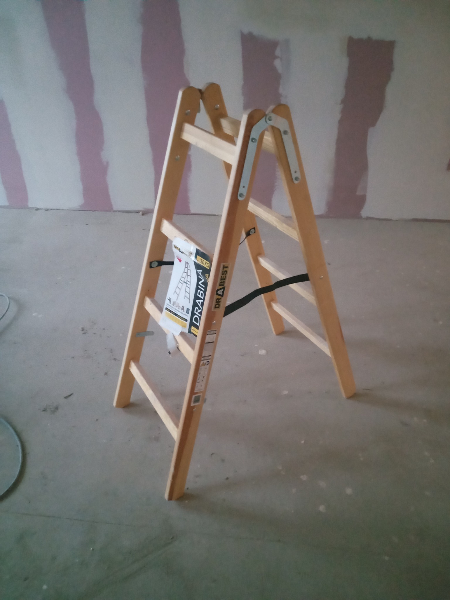 Drewniana drabina rozstawiona w pomieszczeniu z częściowo pomalowanymi ścianami w biało-różowe pasy. Widoczny fragment podłogi i ściany w trakcie prac wykończeniowych.