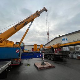 KWP POLAND SPÓŁKA Z OGRANICZONĄ ODPOWIEDZIALNOŚCIĄ - Żuraw samochodowy marki Demag z wysięgnikiem DZWIGAJ.COM podnosi element konstrukcyjny na tle hali przemysłowej i pochmurnego nieba.