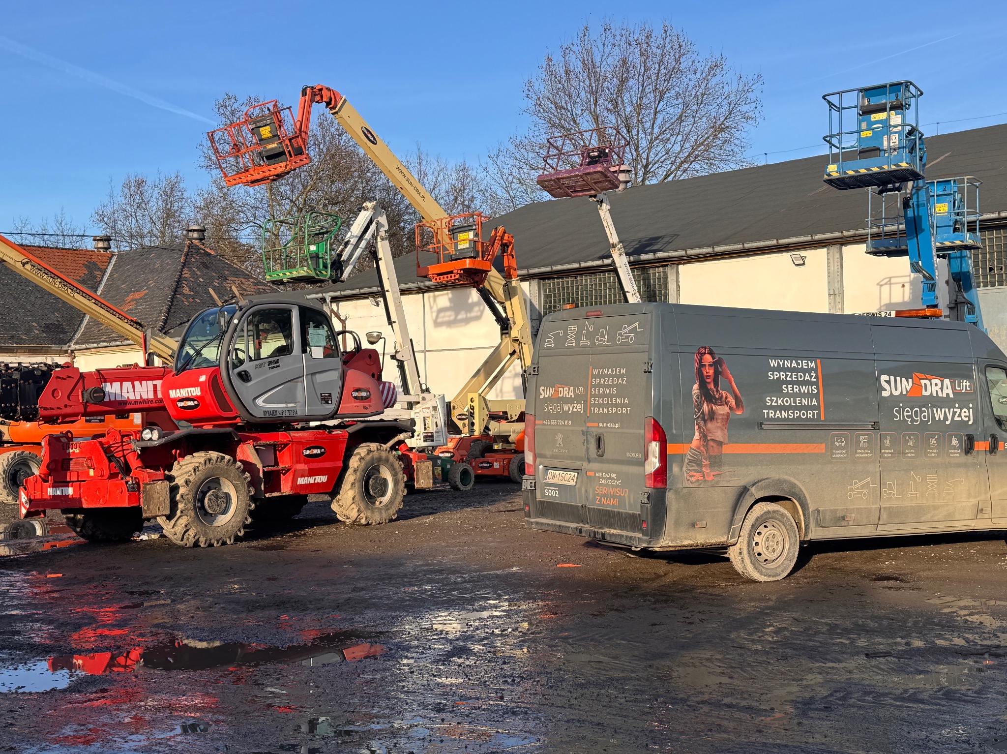 Park maszyn Sun-Dra Lift: podnośniki koszowe, ładowarka teleskopowa Manitou i bus z reklamą usług: wynajem, sprzedaż, serwis, szkolenia i transport. Błotnisty teren.