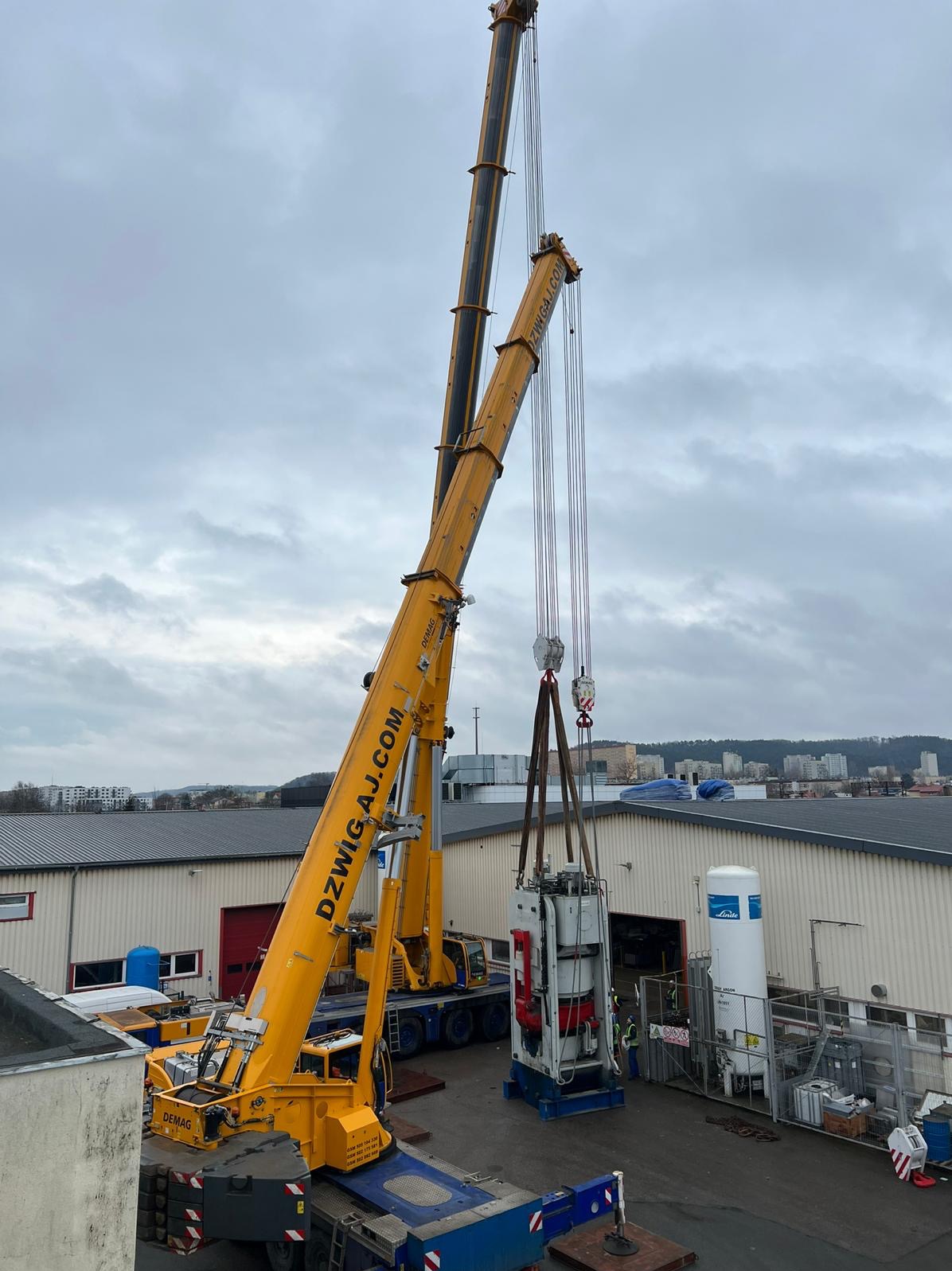 Żółty dźwig Demag podnosi ciężką maszynę na tle budynków i pochmurnego nieba. Widoczne liny, haki i elementy konstrukcyjne dźwigu. Dźwig firmy DZWIG.AJ.COM.