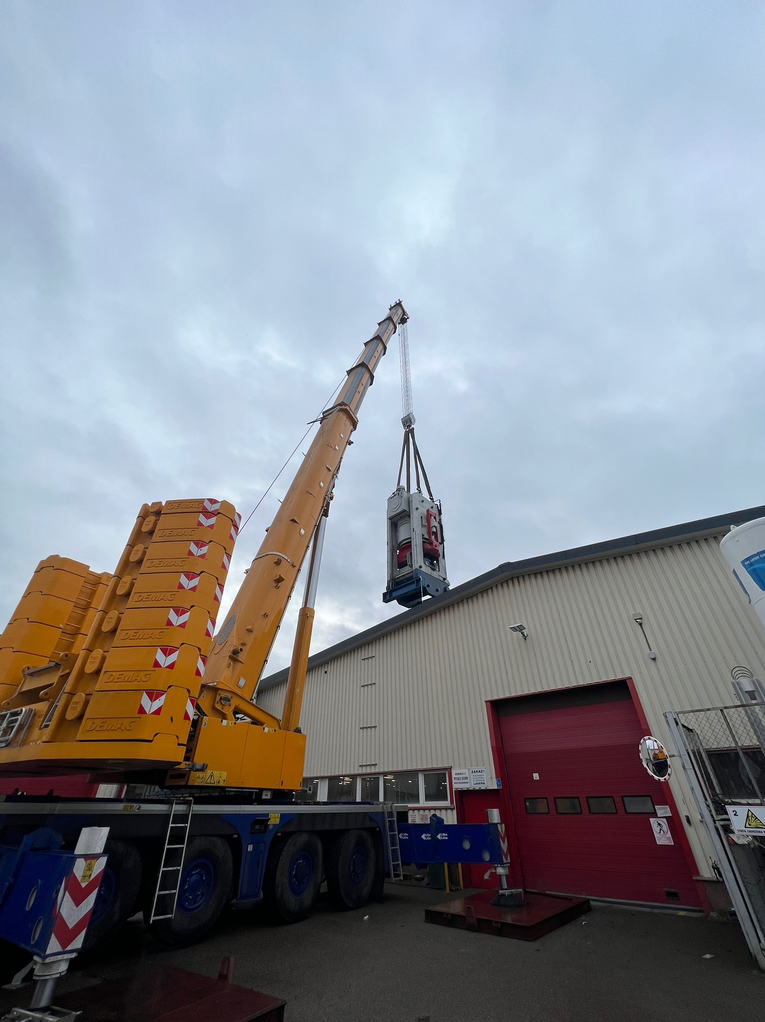 Żuraw Demag podnosi maszynę na tle hali z czerwoną bramą. Widok z dołu, pochmurne niebo. Żuraw z wysięgnikiem teleskopowym i przeciwwagą.