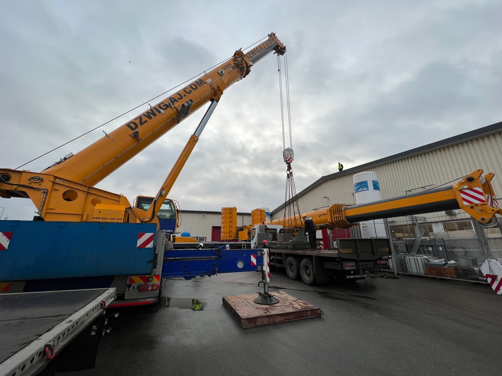 Żuraw samochodowy marki Demag z wysięgnikiem DZWIGAJ.COM podnosi element konstrukcyjny na tle hali przemysłowej i pochmurnego nieba.