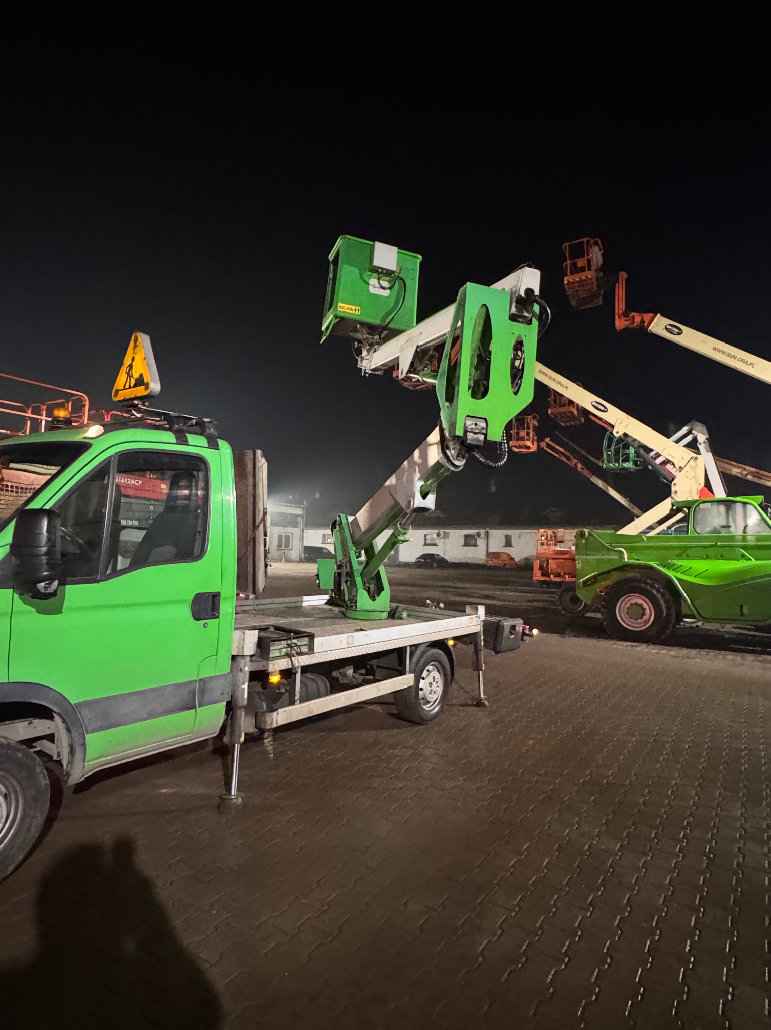 Zielony podnośnik koszowy na samochodzie dostawczym, z wysięgnikiem i koszem. W tle inne podnośniki i budynek. Nocne oświetlenie.