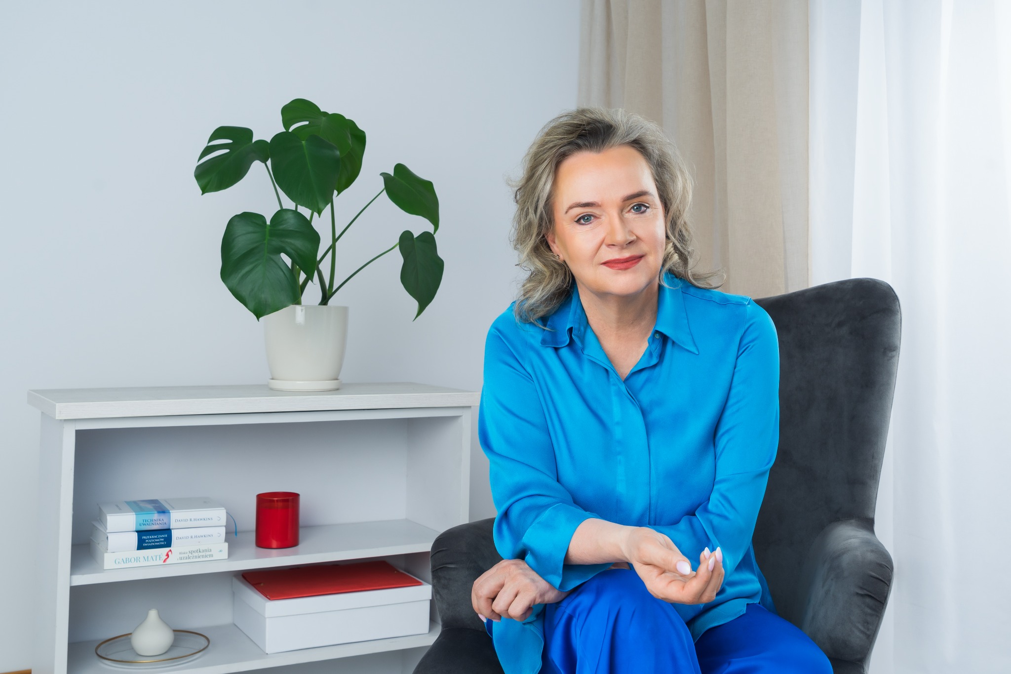 Portret kobiety w błękitnej koszuli siedzącej w fotelu, z rośliną monstera i regałem w tle. Uśmiech, kontakt wzrokowy, jasne wnętrze.