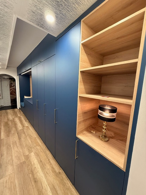 Długa, zabudowana szafa wnękowa w odcieniach granatu z drewnianymi półkami i oświetleniem, dopasowana do wąskiego korytarza z drewnianą podłogą.