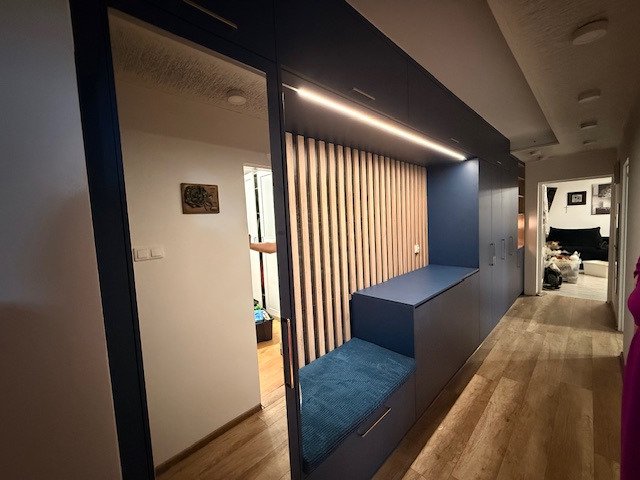 Długa szafa wnękowa w kolorze granatowym z siedziskiem i drewnianymi lamelami, oświetlona listwą LED, w korytarzu z drewnianą podłogą i lustrem.