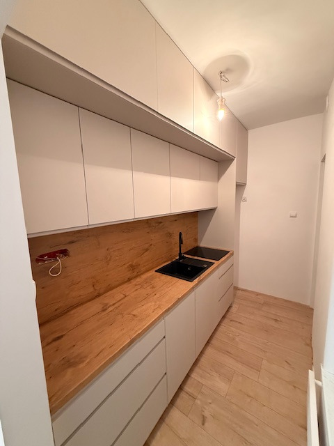 Jasna kuchnia z drewnianym blatem i czarnym zlewem. Górne szafki sięgają sufitu, a podłoga wyłożona jest panelami imitującymi drewno. Minimalistyczny design.