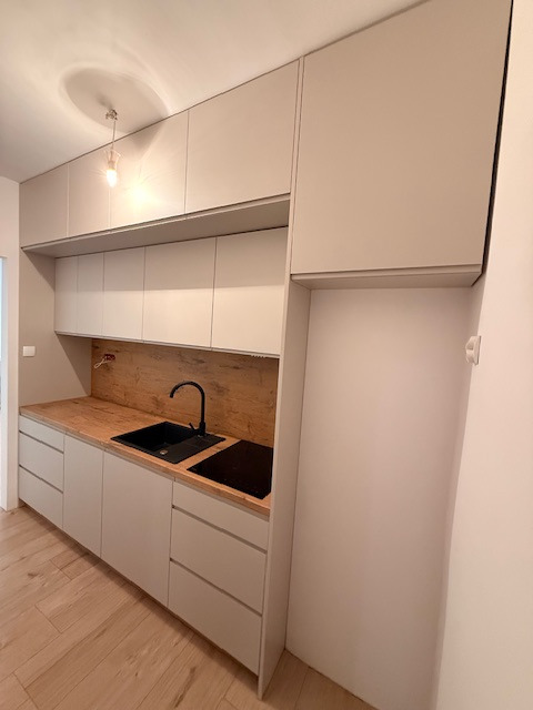 Jasna kuchnia z drewnianym blatem i czarnym zlewem. Górne szafki sięgają sufitu, a dolne mają szuflady. Minimalistyczny design z oświetleniem punktowym.