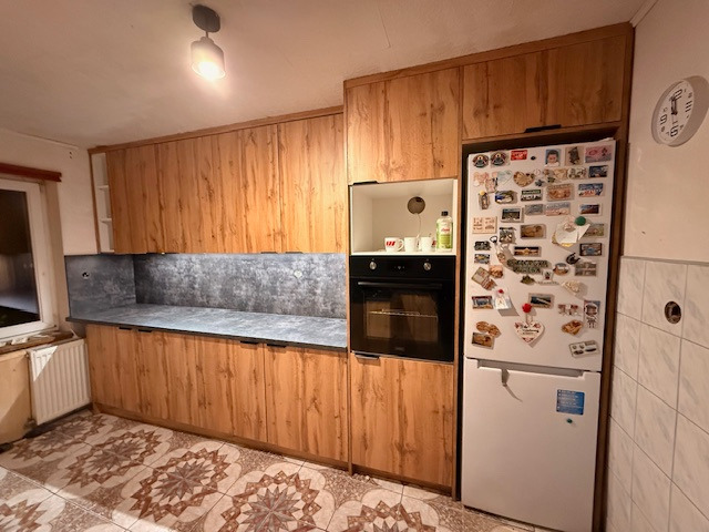 Jasne drewniane meble kuchenne z szarym blatem i lodówką obok piekarnika. Na lodówce magnesy. Na podłodze wzorzyste płytki. Całość w ciepłym oświetleniu.