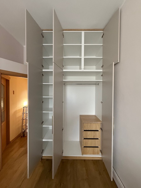 Otwarta szafa wnękowa z szarymi drzwiami i drewnianymi szufladami, widoczne półki i drążek na ubrania. W tle widoczny fragment pokoju z drabiną.