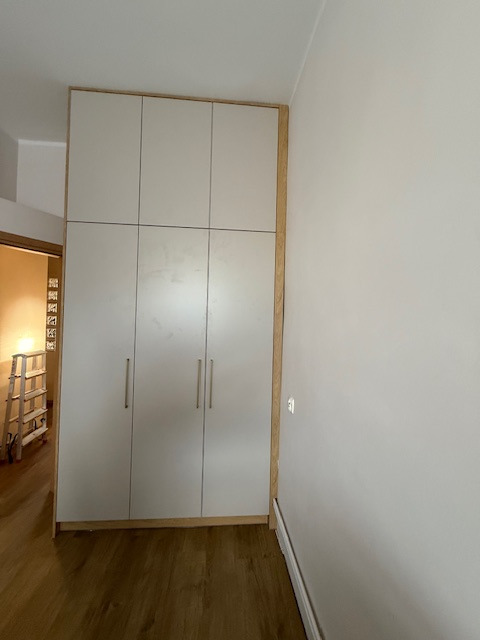 Biała szafa wnękowa z sześcioma drzwiami, drewniane wykończenie, widok z boku, parkiet na podłodze, drabina w tle, minimalistyczny design.