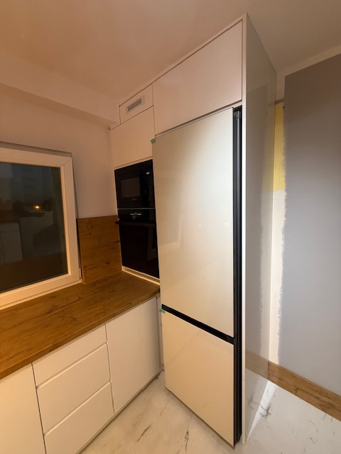 Biała zabudowa kuchenna z drewnianym blatem pod oknem, piekarnikiem i lodówką w zabudowie. Minimalistyczny design, jasne wnętrze, marmurowa podłoga.