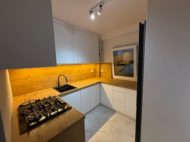 Nowoczesna kuchnia z białymi szafkami i drewnianym blatem, oświetlona ciepłym światłem LED. Czarny zlew i płyta gazowa dodają kontrastu. Widok na okno.