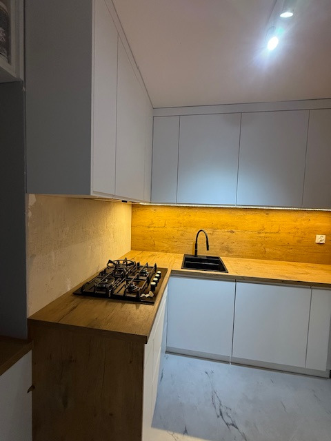 Nowoczesna kuchnia z białymi szafkami i drewnianym blatem, oświetlona listwą LED. Czarny zlew i płyta gazowa dodają kontrastu. Marmurowa podłoga.