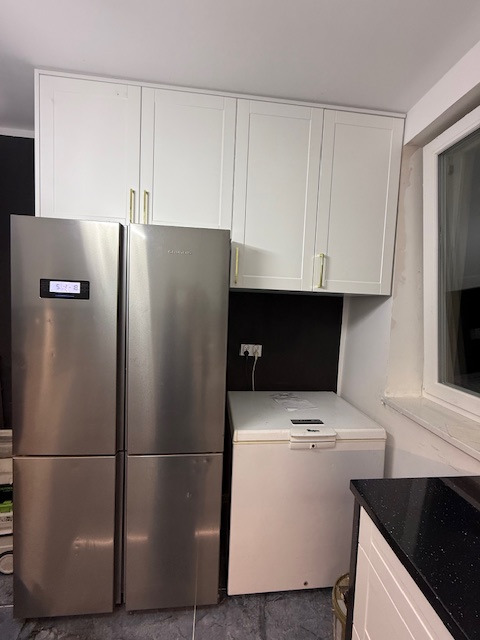 Białe szafki kuchenne z złotymi uchwytami nad dwoma srebrnymi lodówkami i białą zamrażarką skrzyniową, czarny panel ścienny, szara podłoga.