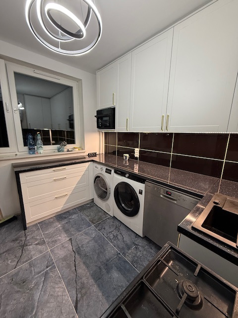 Biała kuchnia z czarnym blatem i sprzętami AGD: pralka, zmywarka. Nad blatem brązowe płytki i szafki wiszące. Na suficie nowoczesna lampa. Marmurowe płytki na podłodze.