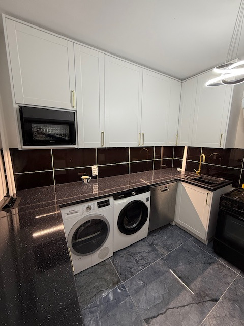 Biała zabudowa kuchenna z czarnym blatem, wkomponowana pralka i suszarka. Złote uchwyty i bateria. Nowoczesne oświetlenie sufitowe. Ciemna glazura.