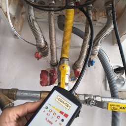 TEMP-EL SERWIS TOMASZ SZABAT - Kontrola szczelności instalacji gazowej detektorem DGN-5B. Widoczne metalowe rury, zawory i przewody. Żółty przewód gazowy. Biała ściana w tle.