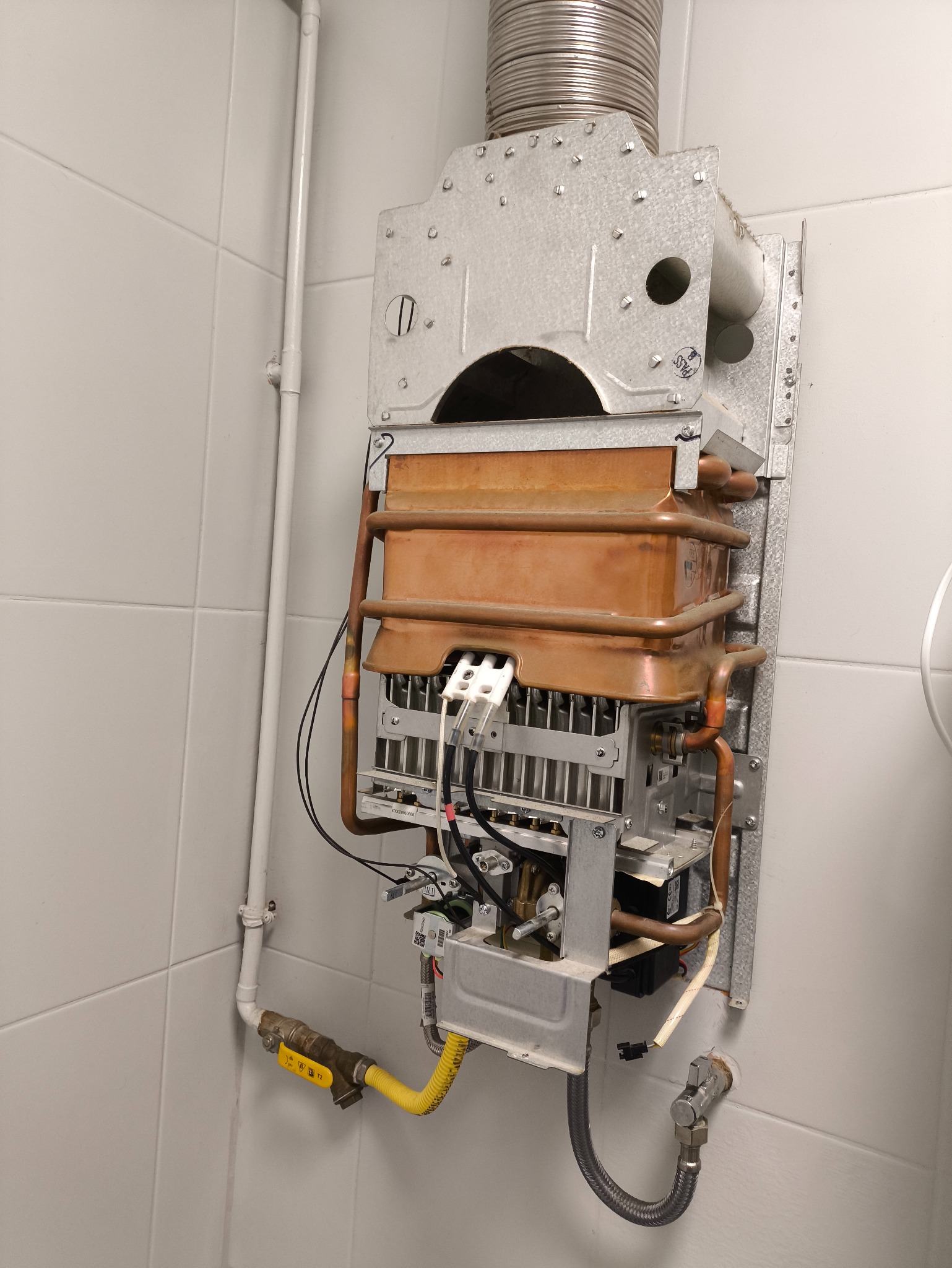 Otwarty piec gazowy z widocznymi elementami wewnętrznymi, rurami miedzianymi i przewodami, zamontowany na białej ścianie z płytek. Widoczny przewód kominowy.