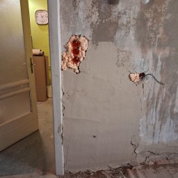 Jonatan Wiśniewski JON-BUD - Ściana w trakcie szpachlowania z widocznymi puszkami elektrycznymi wypełnionymi pianką montażową. Fragment otwartych drzwi i podłogi w tle. Widoczna żarówka na kablu.