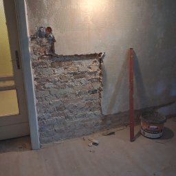 Jonatan Wiśniewski JON-BUD - Odsłonięta cegła przy drzwiach, fragment ściany z instalacją elektryczną, poziomica i wiadro z zaprawą. Przygotowanie do prac wykończeniowych.