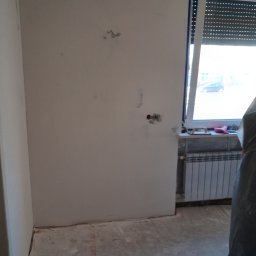 Jonatan Wiśniewski JON-BUD - Pokój w trakcie remontu: ściany po szpachlowaniu, widoczne kable, grzejnik pod oknem. Podłoga przygotowana do dalszych prac. Częściowo zakryty element w folii ochronnej.