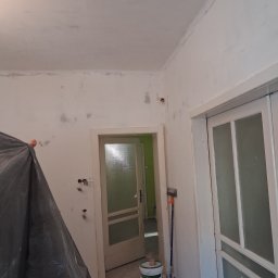 Jonatan Wiśniewski JON-BUD - Pokój w trakcie szpachlowania: ściany z widocznymi łatami szpachli, zakryte meble, otwarte drzwi do kolejnego pomieszczenia, wiadro z farbą i wałek malarski.