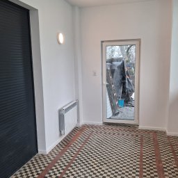 Jonatan Wiśniewski JON-BUD - Białe ściany po malowaniu, czarne drzwi z żaluzjami, wzorzysta podłoga z płytek i grzejnik w odnowionym wnętrzu. Widok na zewnątrz przez przeszklone drzwi.