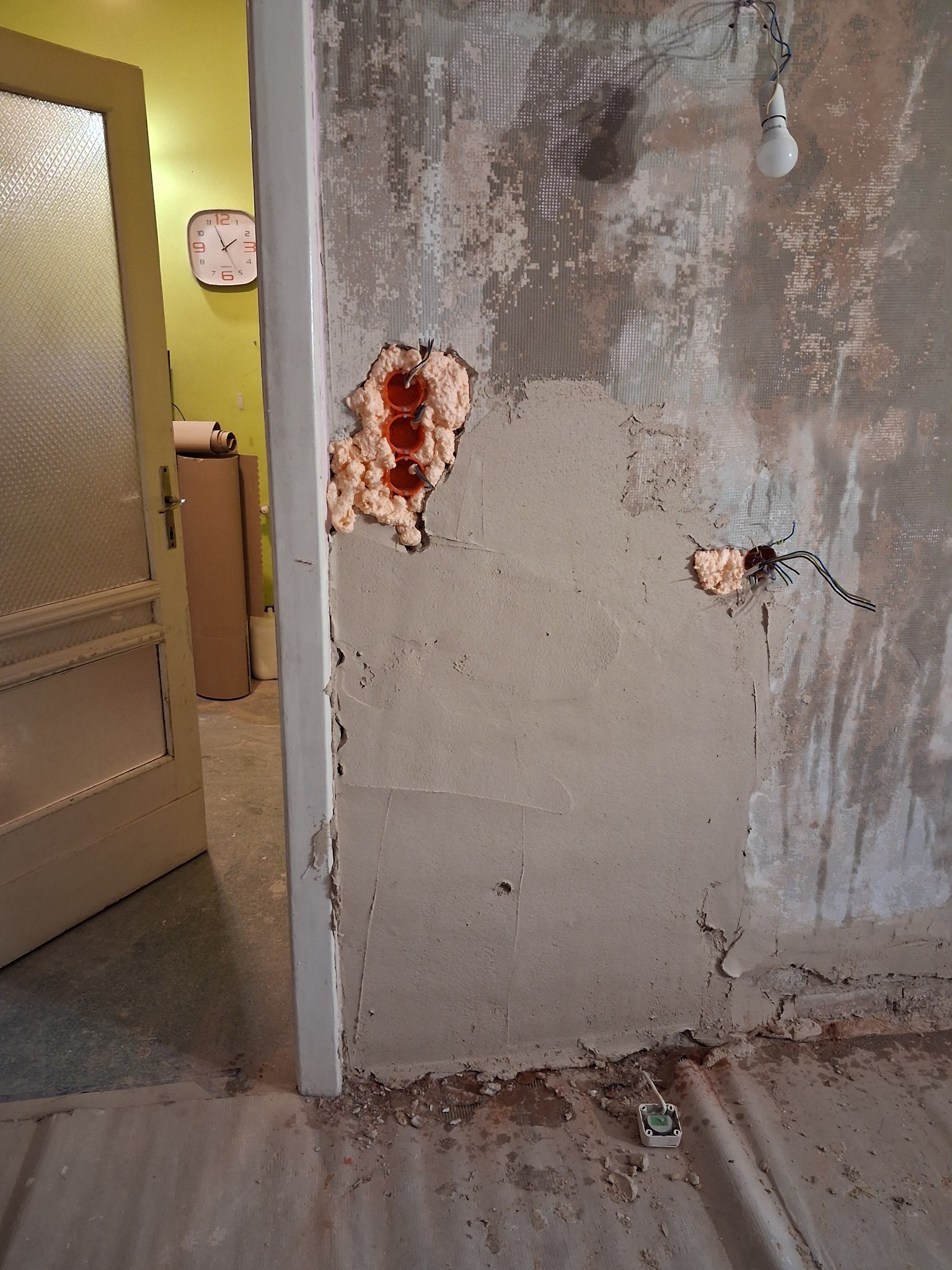 Ściana w trakcie szpachlowania z widocznymi puszkami elektrycznymi wypełnionymi pianką montażową. Fragment otwartych drzwi i podłogi w tle. Widoczna żarówka na kablu.