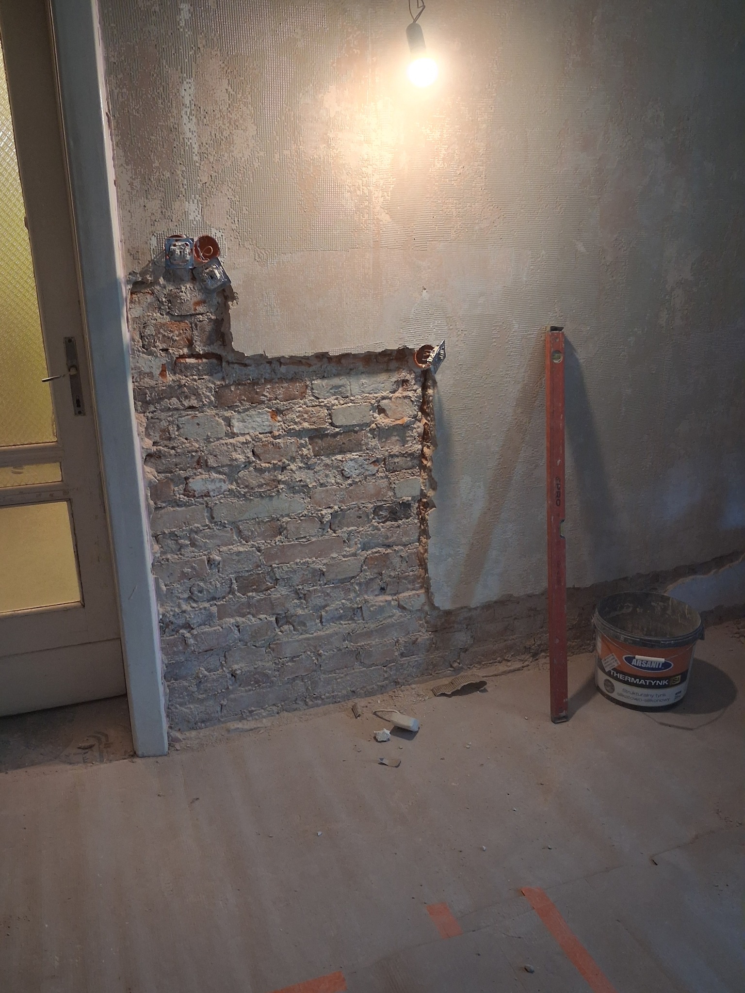 Odsłonięta cegła przy drzwiach, fragment ściany z instalacją elektryczną, poziomica i wiadro z zaprawą. Przygotowanie do prac wykończeniowych.