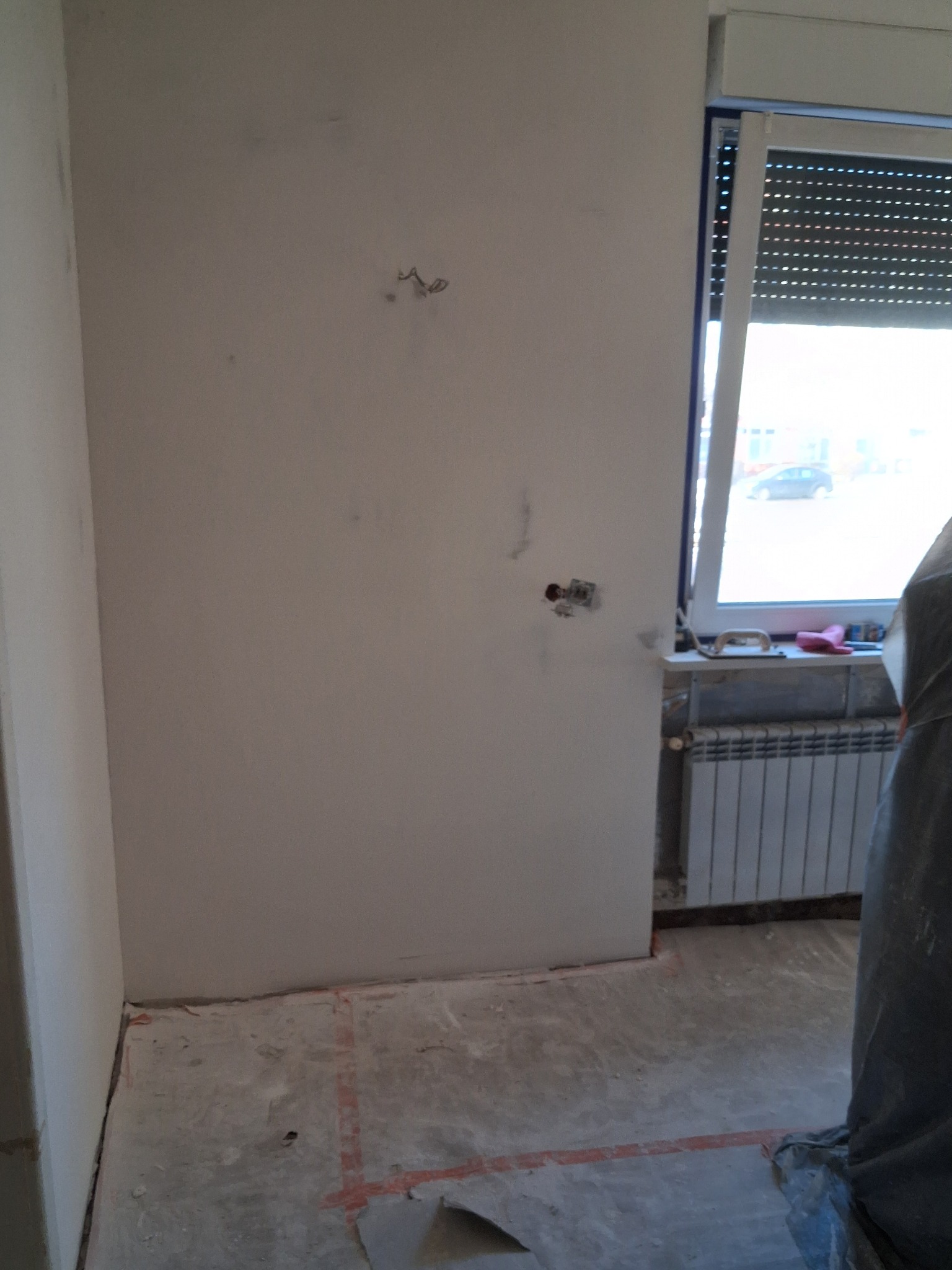 Pokój w trakcie remontu: ściany po szpachlowaniu, widoczne kable, grzejnik pod oknem. Podłoga przygotowana do dalszych prac. Częściowo zakryty element w folii ochronnej.