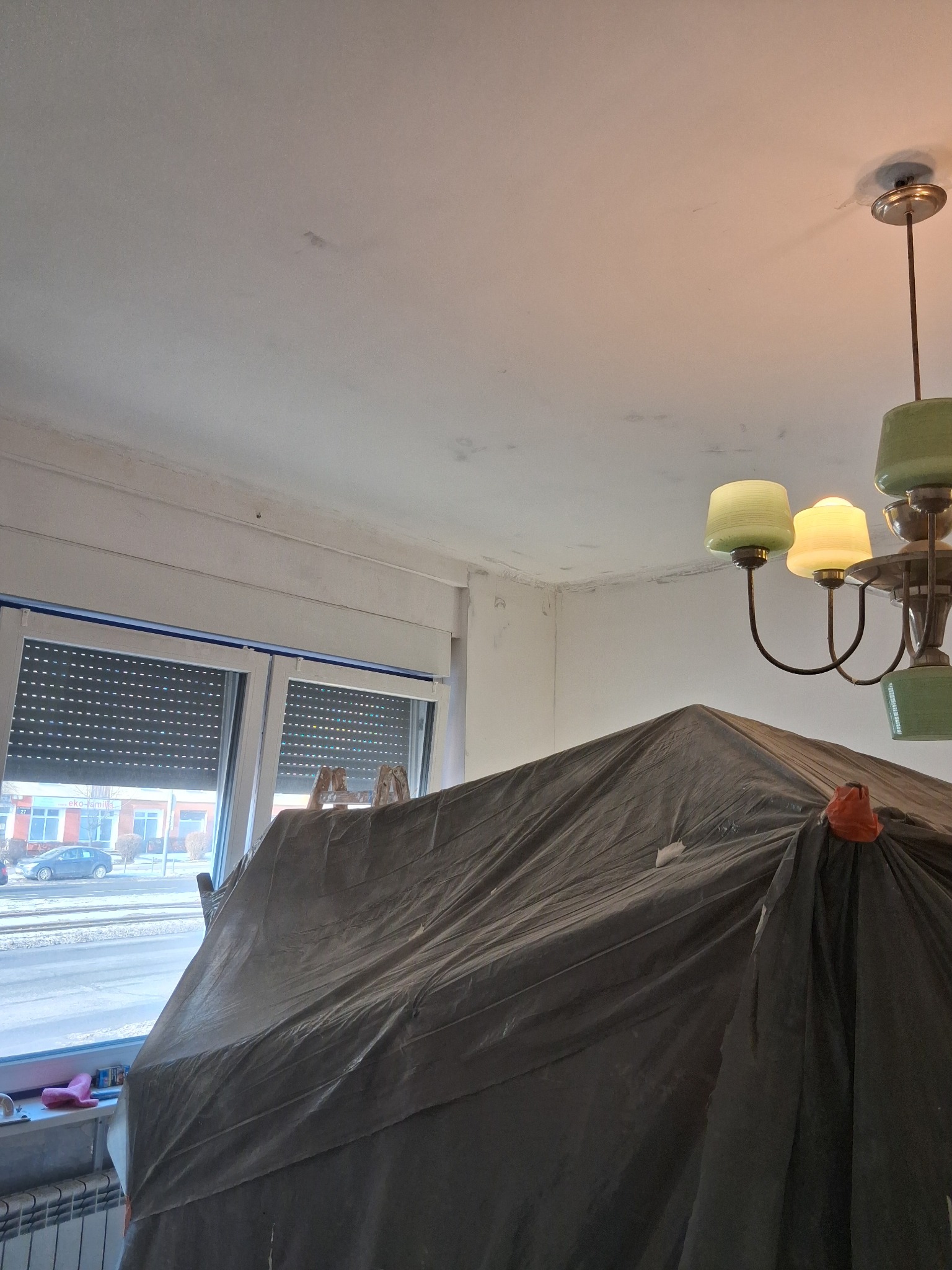 Przygotowanie do malowania: pokój zabezpieczony folią malarską, widoczne ślady szpachlowania na suficie i ścianach, drabina oparta o okno, żyrandol w tle.