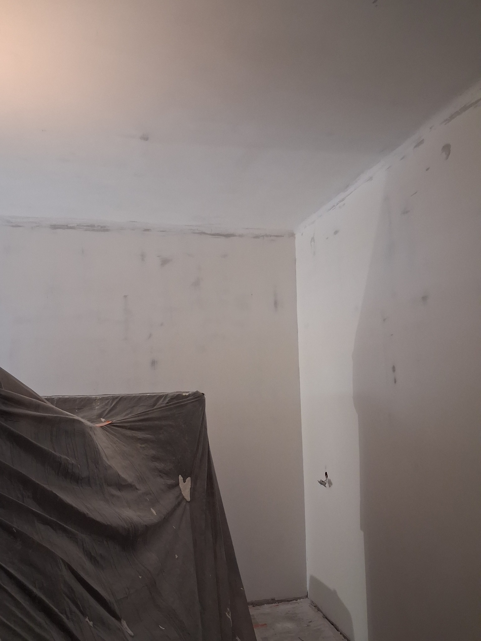 Przygotowanie do malowania: Pokój ze ścianami po szpachlowaniu, zabezpieczony mebel pod szarą folią malarską, widoczne ślady po szpachli na ścianach i suficie.